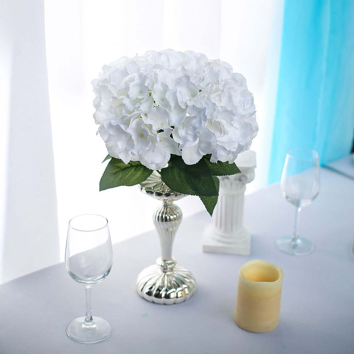 White hydrangea flower arrangement in silver vase on a table with glassware and candle.
