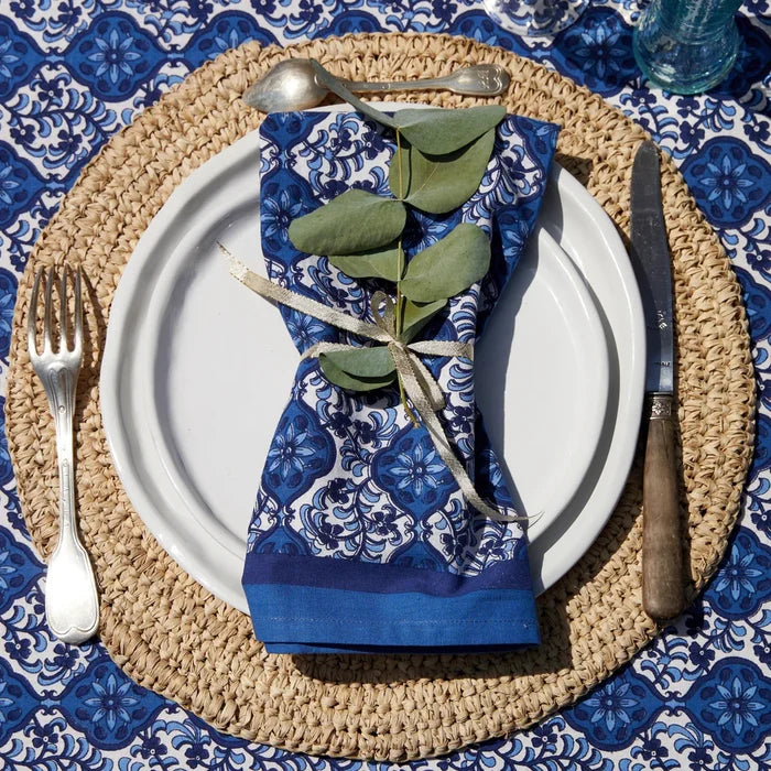 A styled table setting featuring a blue and white patterned cloth napkin tied with a twine bow and green eucalyptus leaves, arranged on a white plate over a woven beige placemat. Silver-toned cutlery and a blue patterned tablecloth are visible in the setting.