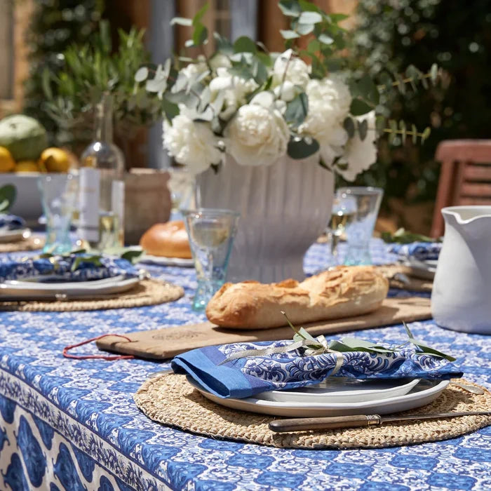 A beautifully styled outdoor dining table set with a blue and white patterned tablecloth, woven placemats, white plates, blue patterned napkins, a loaf of bread, and a large white floral centerpiece in a vase.
