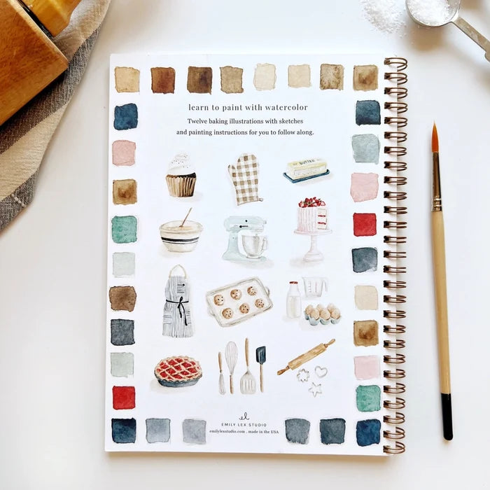 Spiral bound watercolor workbook with paintings of baking related items sitting next to a paintbrush