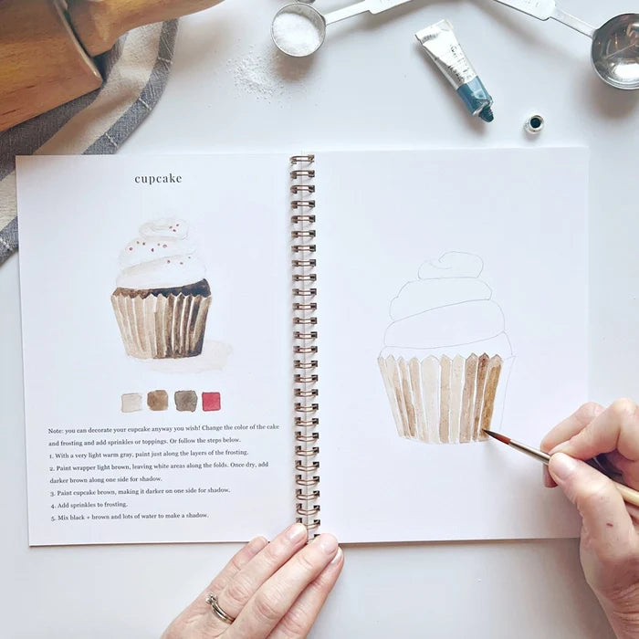 Open book with cupcake illustration and hands painting a cupcake
