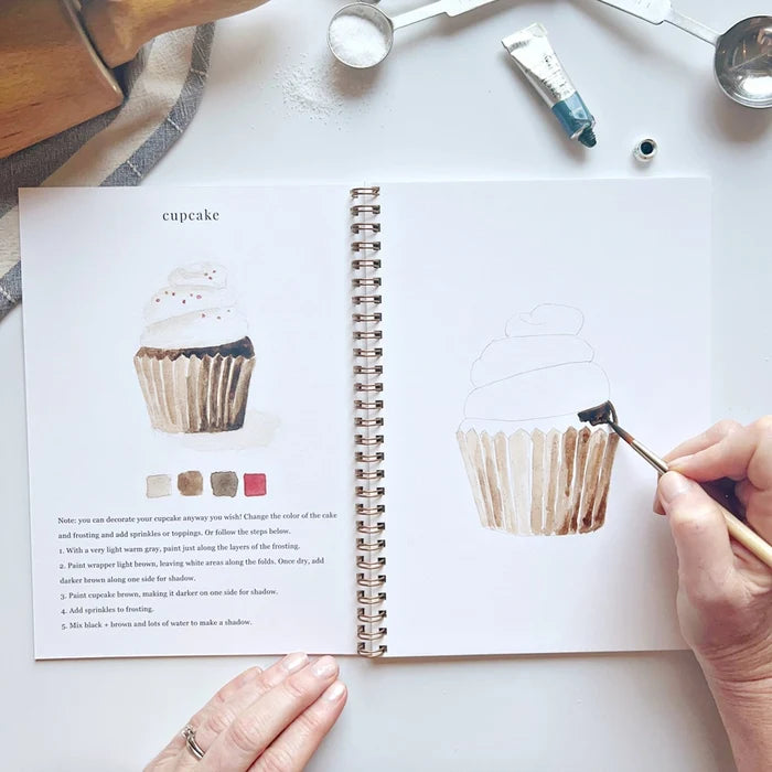 Open book with drawing of a cupcake and hand holding a brush
