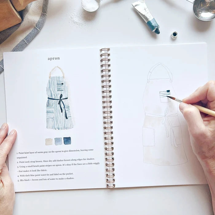 Person holding a paintbrush over an open book with apron painting