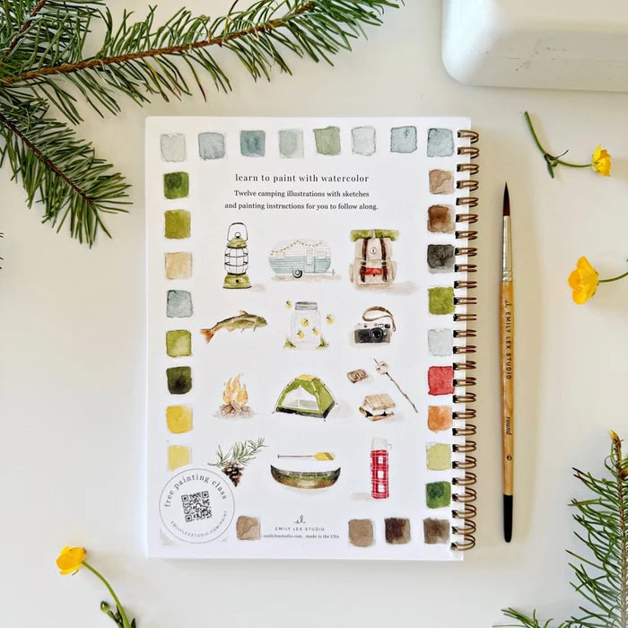 Notebook with watercolor camping themed  illustrations on a white surface with a paintbrush