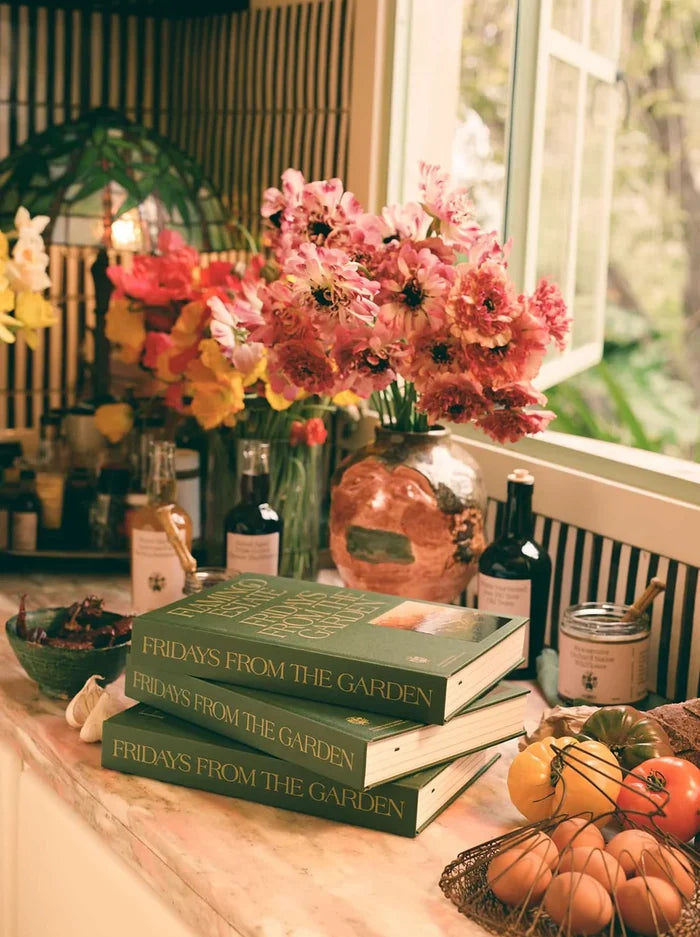 Three stacked green books titled 'FRIDAYS FROM THE GARDEN' sit on a windowsill surrounded by pink flowers, bottles, eggs, and autumn vegetables in a warm, sunlit indoor setting.