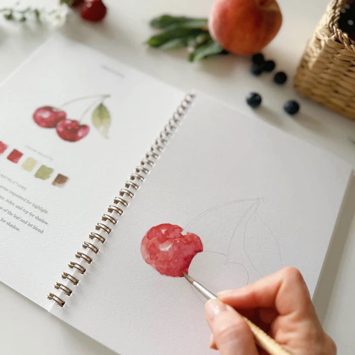 Person painting a cherry on a sketchbook with fruit and a basket in the background
