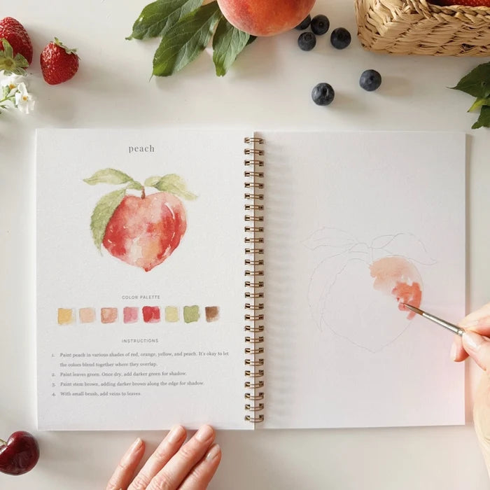 Open art book with a peach illustration and color swatches, surrounded by fruits