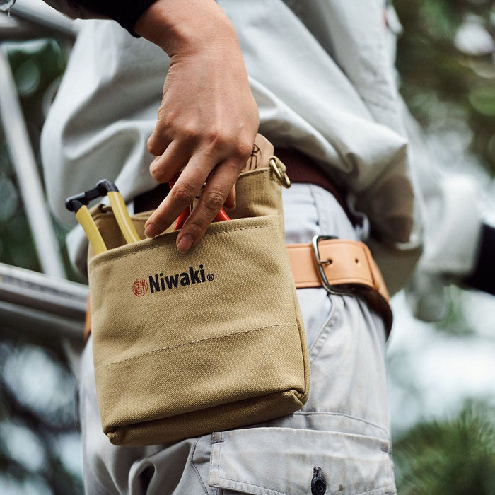 Person holding a tan tool pouch with 'Niwaki' branding outdoors.