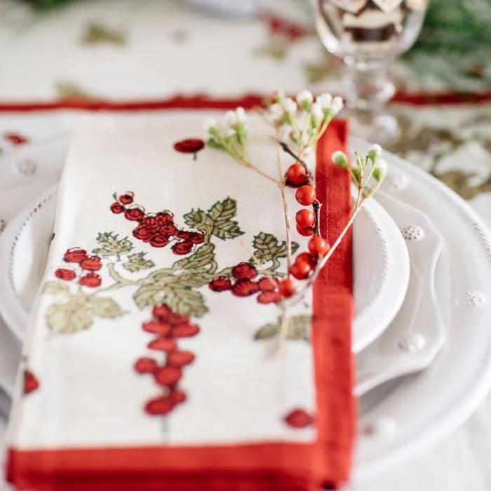 A close-up of a white table setting featuring a folded napkin with a red berry and green leaf pattern, a red ribbon border, and a sprig of red berries on top, layered over white plates.