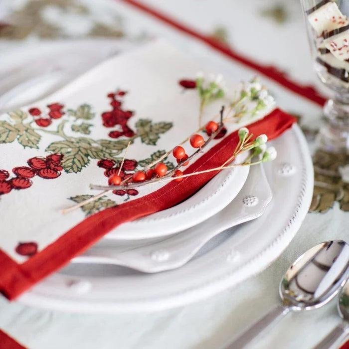 A close-up of a christmas table setting featuring a white ceramic plate stack with a cream napkin printed with red berries and green leaves, accented by a red ribbon and small decorative berry sprigs.