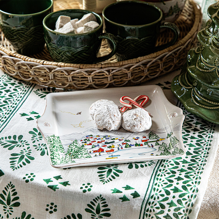 A white tray painted with a winter town scene sitting on a table with powdered sugar covered cookies, a wicker basket with green mugs filled with homemade marshmallows