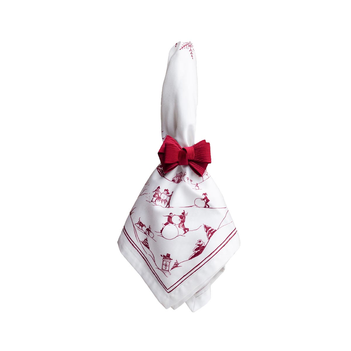 A white napkin with a red toile-style print of sleighs and figures, tied with a red bow napkin ring
