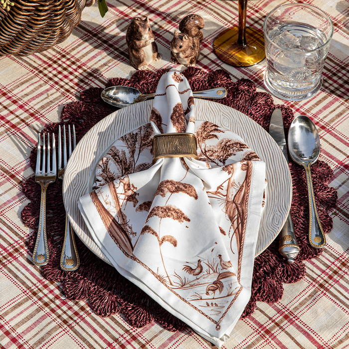 A styled table setting featuring a white napkin with a brown botanical and bird pattern, folded on top of a white plate and secured with a gold ring. The setting is placed on a dark red textured placemat over a red and beige plaid tablecloth, with silver cutlery, a glass of water, and small decorative animal salt and pepper shakers in the background.