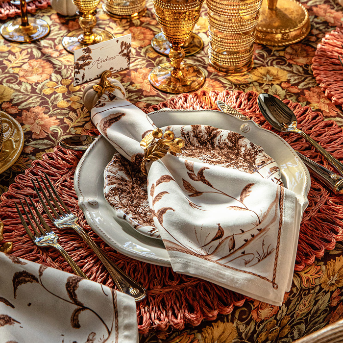 A styled table setting featuring a white linen napkin with a brown botanical leaf pattern, folded and tied with a gold blueberry ring, resting on a white charger plate over a rust-colored placemat. Matching napkins and gold tableware are arranged around it on a patterned tablecloth.