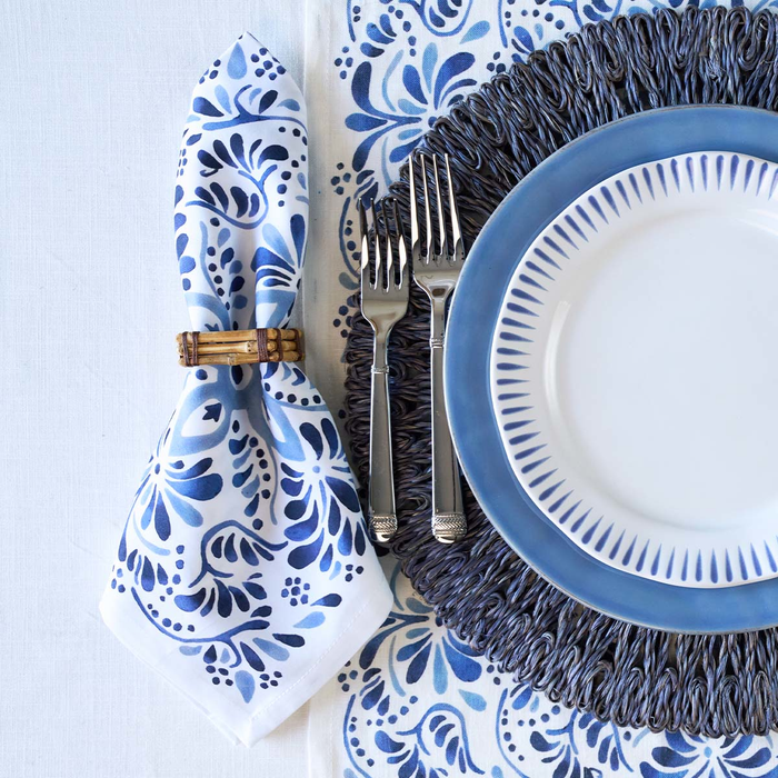 A styled table setting featuring a blue and white floral patterned napkin tied with a bamboo ring, a blue and white plate, silver forks, and a dark blue textured charger on a white tablecloth.