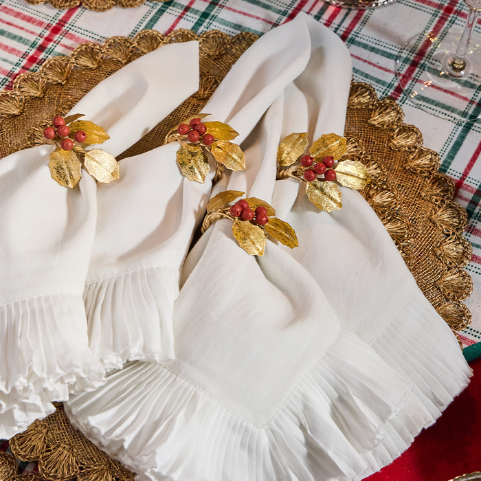 White ruffled napkins with gold holly leaf and red berry napkin rings, arranged on a woven placemat over a plaid tablecloth.