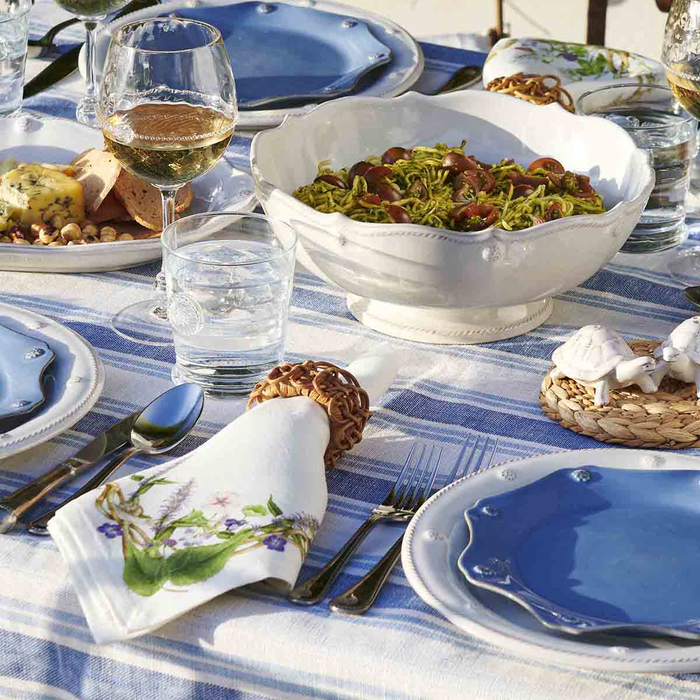 A styled dining table setting featuring a white scalloped bowl, blue and white plates, a white napkin with floral print, and a striped blue-and-white tablecloth.