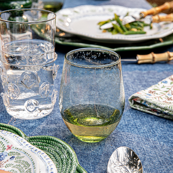 Green tinted stemless wineglass  filled with white wine on a table with plates, glasses, and cutlery.