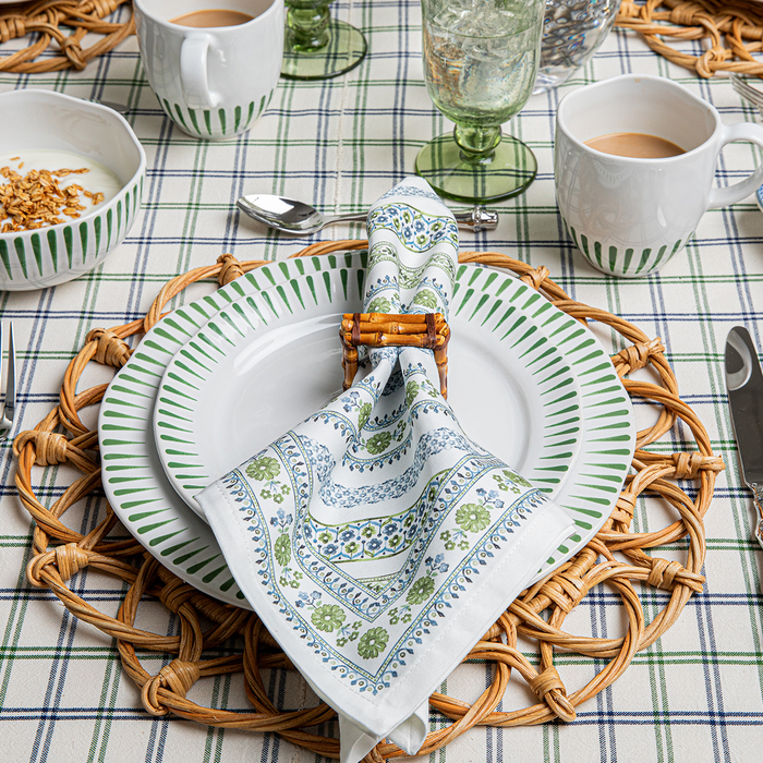 Table setting with white plates, green rimmed cups, and a green and blue floral and stripe patterned napkin on a checkered tablecloth.