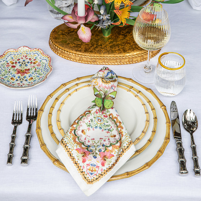 A styled table setting featuring a white tablecloth, a bamboo-rimmed plates, a white napkin with a colorful floral and butterfly pattern, silver bamboo shaped cutlery, a glass of white wine, and a floral centerpiece.