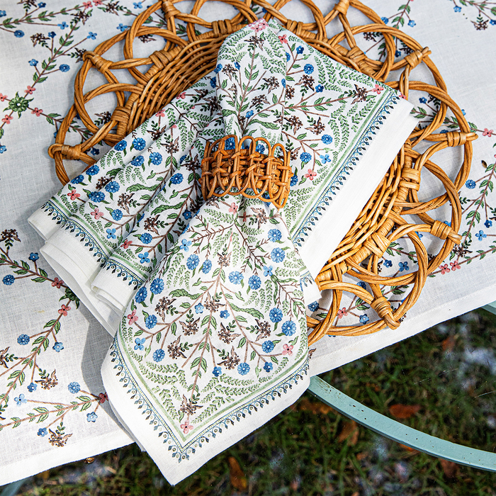 A stack of floral napkins with the top one tied with a wicker napkin ring on a wicker placemat and floral printed tablecloth