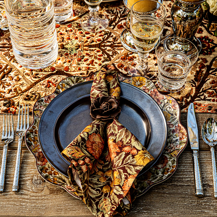 An autumn-themed table setting featuring a dark slate  blue plate on an autumnal charger with a scalloped edge. The place setting includes a folded floral napkin in warm fall colors, silver flatware, and drinking glasses on a rustic wooden table.
