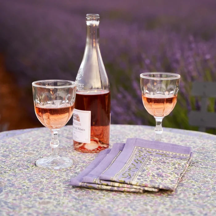 A bottle and two stemmed glasses of rosé wine on a table beside folded patterned napkins, with a blurred lavender field in the background.
