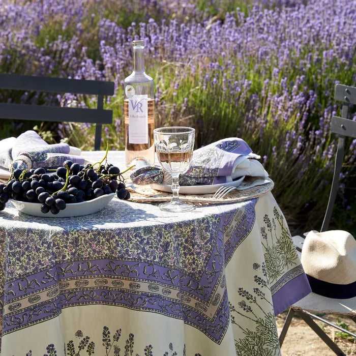 A round table set outdoors in a lavender field with a cream tablecloth featuring a purple botanical border pattern. The table is styled with a bottle of wine, a filled wine glass, plates, folded napkins, a bowl of dark grapes, and chairs are visible in the background.
