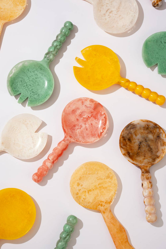 A collection of colorful, handcrafted soap or resin spoons and small dishes scattered on a clean white background. The items feature marbled patterns in yellow, green, pink, brown, and cream.