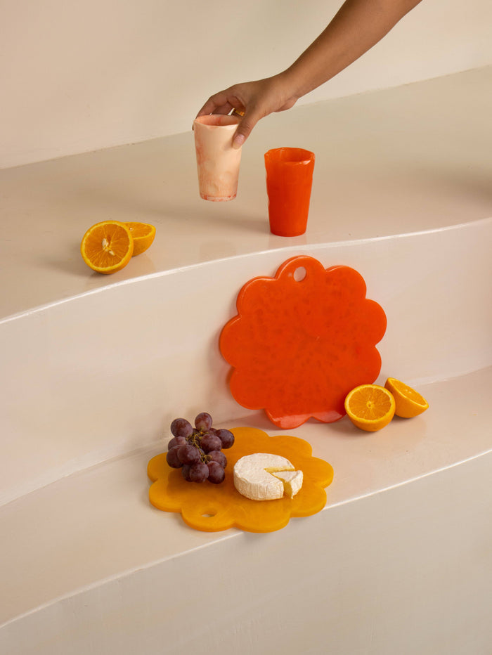 A hand holds a translucent peach tumbler above a bright orange cup on a cream-colored tiered platform. Below, a yellow scalloped cheese board holds grapes and a cheese slice, with an orange scalloped board standing behind it, alongside orange slices.
