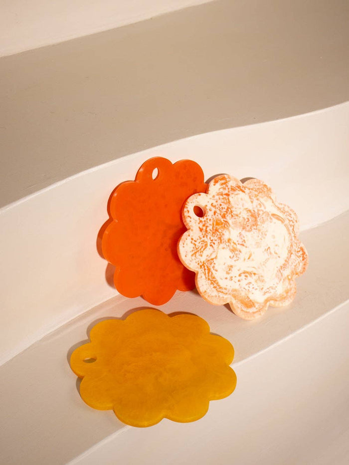 Three scalloped flower-shaped bath or body soaks in orange, mustard yellow, and peachy white, arranged on a white curved surface.