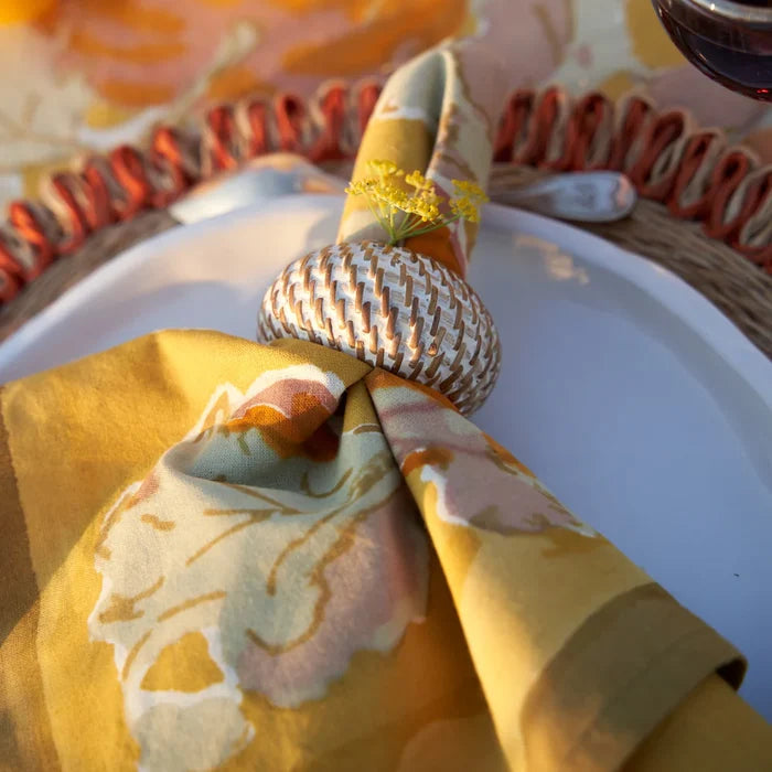 Decorative napkin with a fall leaf design on a white plate, set against a blurred background of a table setting.