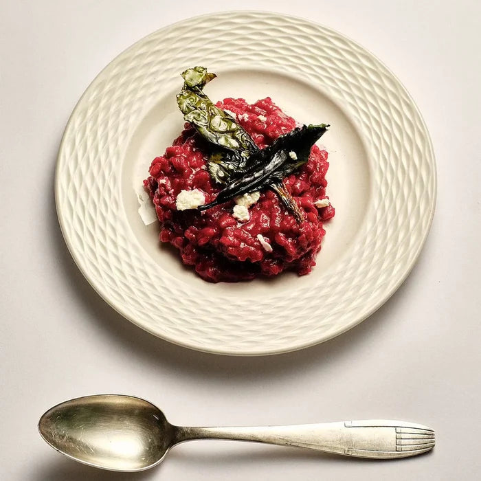 A plated food dish showing a mound of dark red risotto with a leaf garnish and small white crumbles, served on a textured off-white plate with a silver spoon below on a light gray surface.