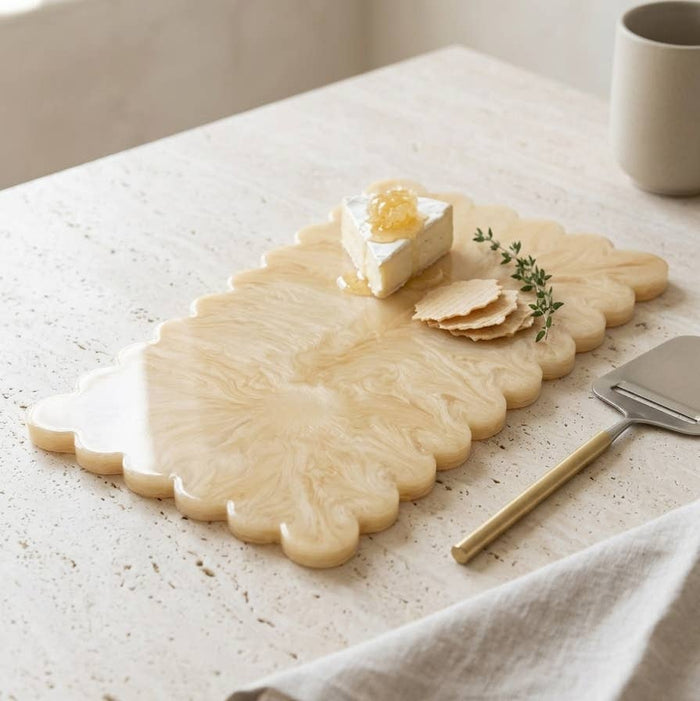 A scalloped cream-colored stone serving board with marbled swirls, styled on a speckled stone table with cheese, crackers, a small sprig of herbs, and a cheese knife.