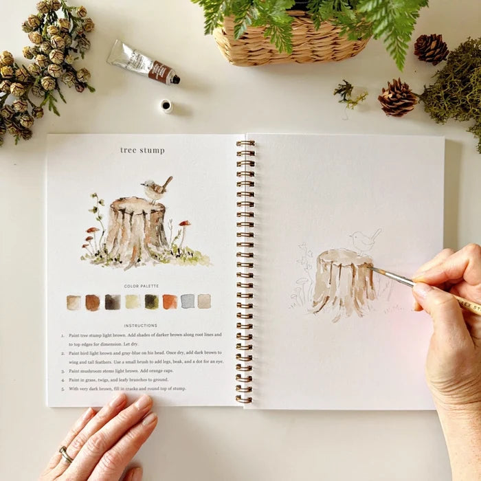 Artistic book with nature-themed illustrations and a hand holding a paintbrush.